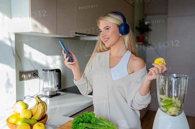 Smiling woman wearing headphones and holding a phone, preparing a healthy smoothie with fresh ingredients in her kitchen, enjoying music and a healthy lifestyle