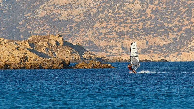 A windsurfer sails near rocks in the blue sea, windsurfer, Meltemi windsurfing spot, Devils Bay, Paralia Vatha, Vatha Beach, south-east coast, Karpathos, Dodecanese, Greek Islands, Greece