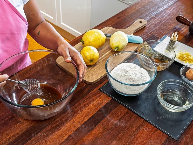 A woman in a pink apron whisks ingredients in a glass bowl on a wooden counter. Nearby, lemons and baking essentials are neatly arranged, ready for making lemon cookies