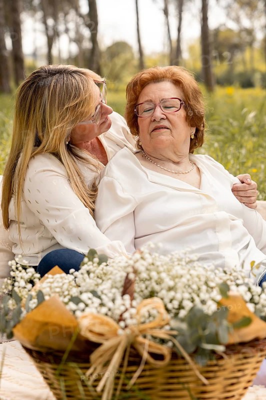 A heartfelt moment between mother and daughter, sharing smiles and love while sitting together in a lush, serene outdoor setting. A basket of flowers adds a delicate touch
