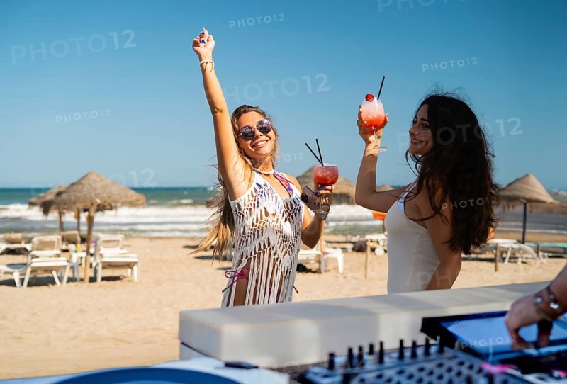 Two friends celebrate summer on a sandy beach, raising their colorful cocktails in the air. Straw umbrellas dot the background, while the waves gently crash nearby, creating a lively atmosphere