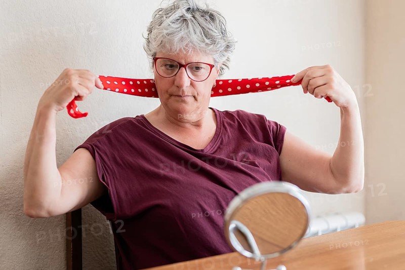 A senior woman with short grey hair and glasses ties a red polka dot scarf around her neck. She sits at a wooden table with a round mirror, focused on her reflection