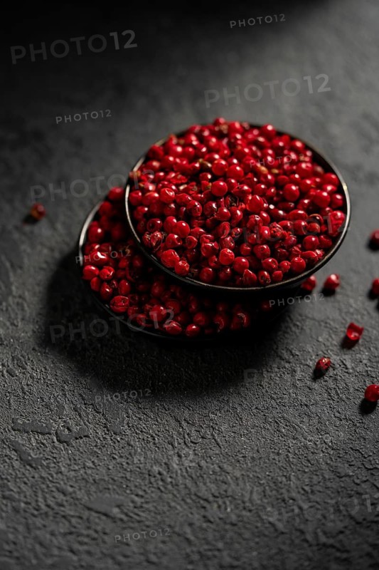 From above, a striking image showing a bowl filled with vibrant red peppercorns placed on a textured slate background. The dark hues contrast the vivid red of the peppercorns, emphasizing their color and texture