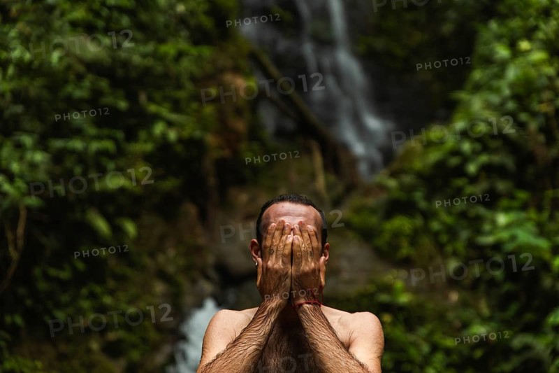 Unrecognizable man covering his face with hands, set against a stunning waterfall in the Choco Andino rainforest of Ecuador. The image captures a sense of mystery and exploration amidst lush tropical landscapes