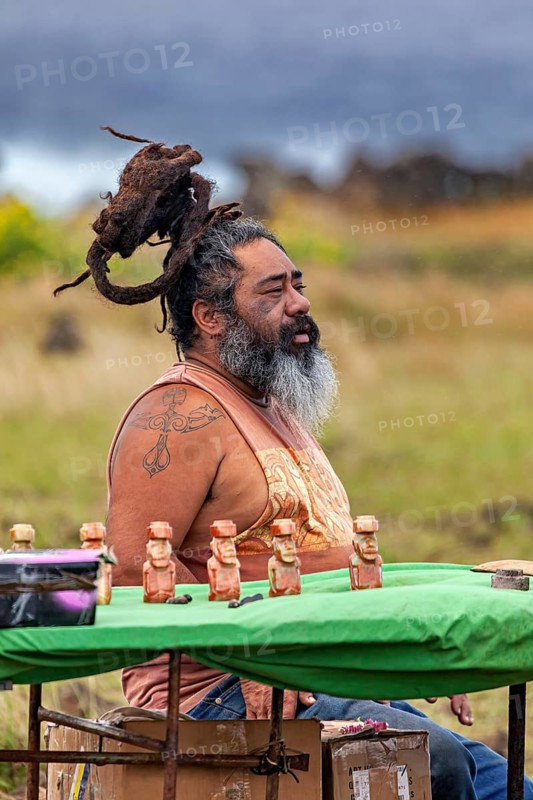 A man with dreadlocks and beard sits outside at a table with small statuettes in a meadow, traditional inhabitant of Easter Island in the Pacific Ocean