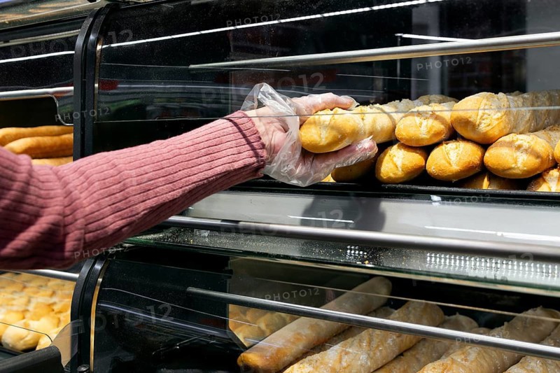 A woman wearing a glove selects fresh bread rolls from a display case at a bakery of a supermarket. The rolls are neatly arranged in rows, showcasing a variety of baked goods