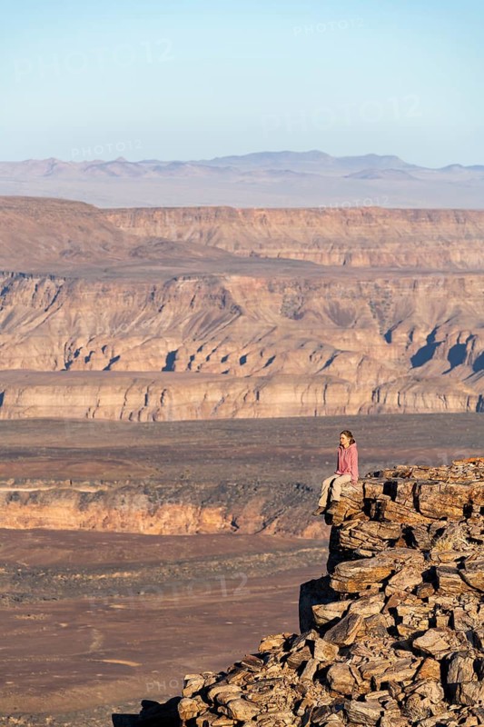 A woman sits on a rock at the edge of the precipice in front of the gorge landscape of the Fish River Canyon, Fish River Gorge, Namibia