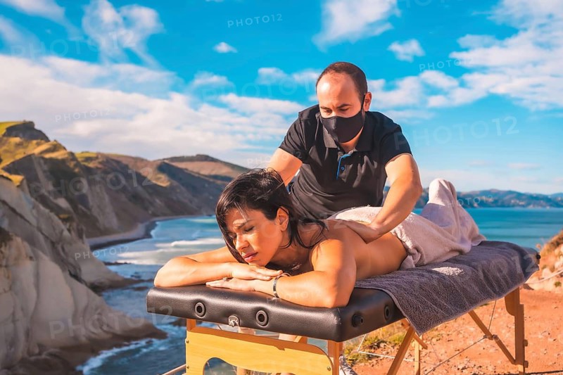 A young man performing a massage in nature on the coast near the sea, a dream come true, masseuse with face mask in the coronavirus pandemic