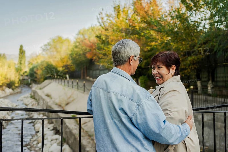 A joyful elderly couple shares a tender moment on a riverside bridge amidst vibrant autumn foliage. They embrace warmly, celebrating love and companionship