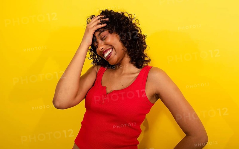 A woman in a vibrant red tank top laughs joyfully, hand on her head, against a bright yellow background. Her curly hair and radiant smile create a cheerful, lively atmosphere