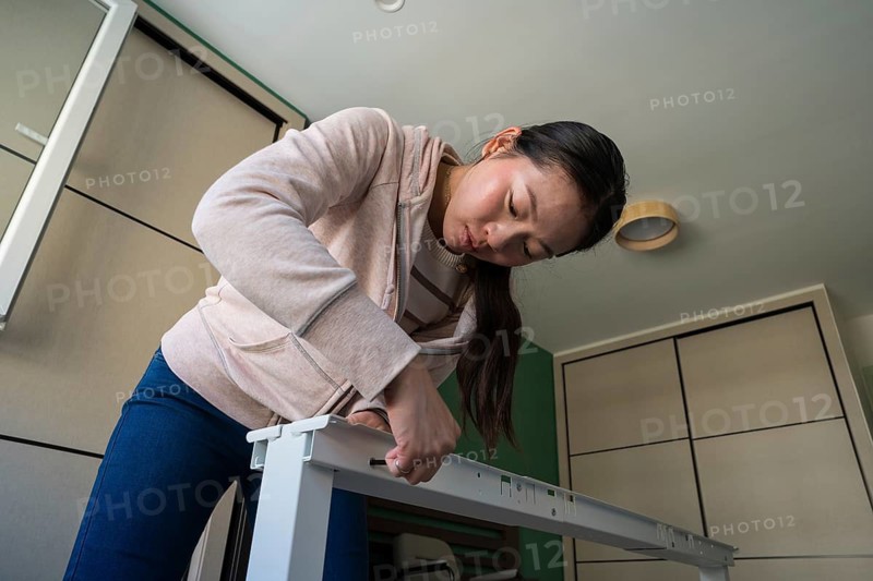 A young Asian woman concentrates as she assembles a white desk at home. Engaged in a DIY furniture project, she carefully follows the instructions, showcasing focus and skill