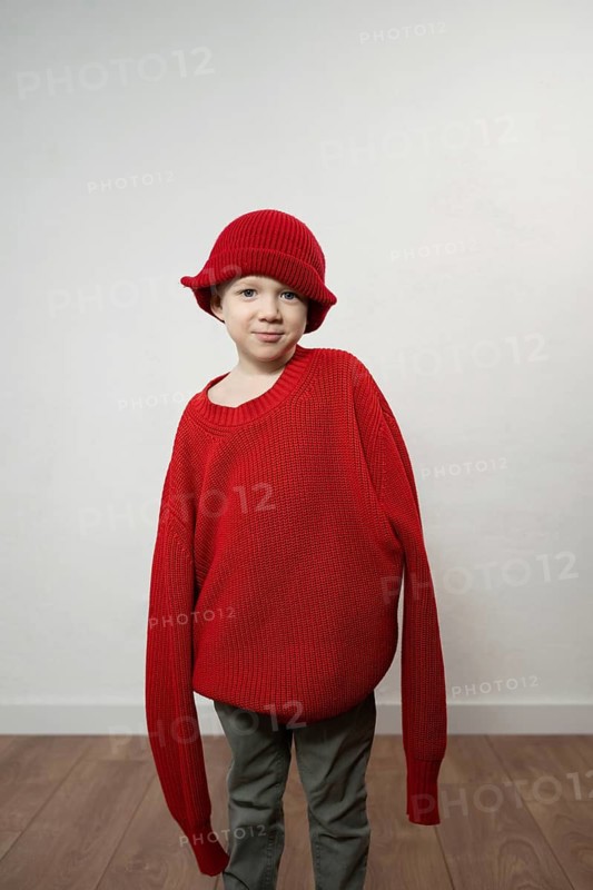 A young child playfully models a cozy oversized red sweater and matching knit hat. The bright red attire contrasts with a simple white background