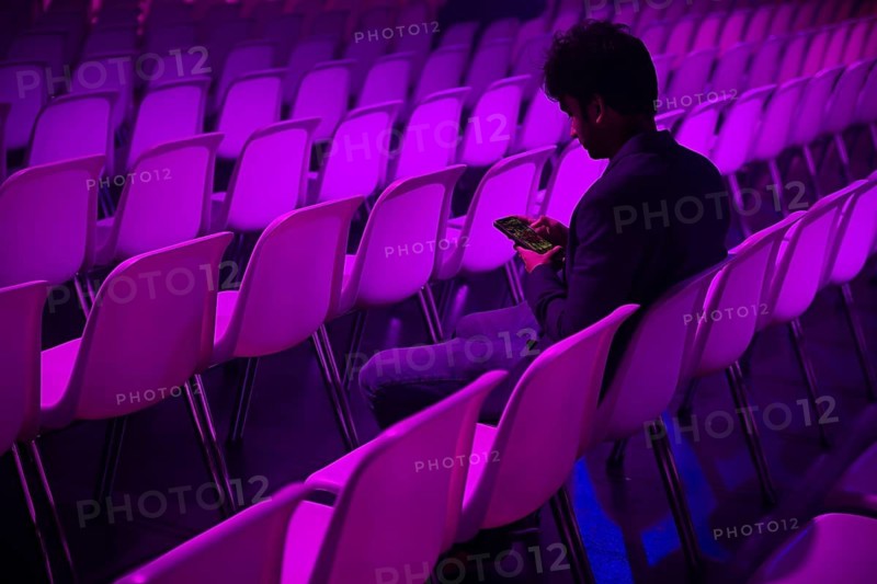 A indian man sits in a purple lit auditorium at a conference of AI in Amsterdam, quietly engaging with their smartphone amidst rows of empty white chairs