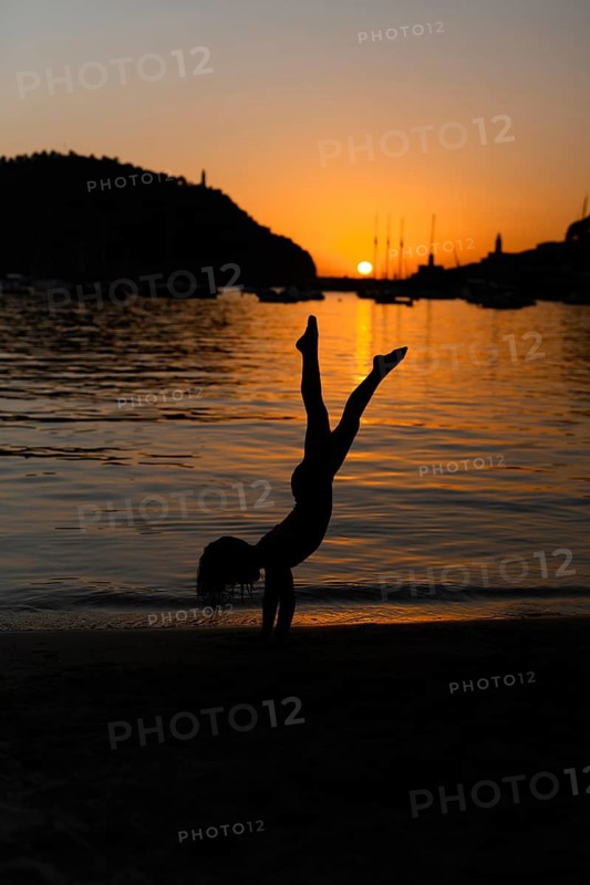 A silhouette of a child doing a handstand on a sandy beach at sunset in Soller, Majorca. The serene waters and a picturesque sunset create a peaceful backdrop for playful energy