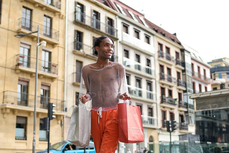 A smiling man holds shopping bags in an urban setting, showcasing confidence and style in a mesh top and bright pants, embodying LGBTQIA+ pride and self-expression