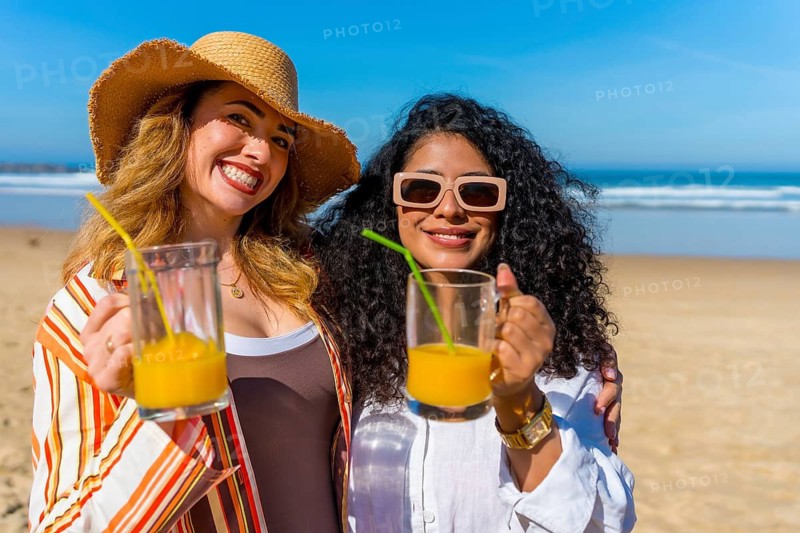 Two smiling women posing for the camera, standing together on a sandy beach holding refreshing orange beverages with the ocean and blue sky in the background, enjoying their summer holiday