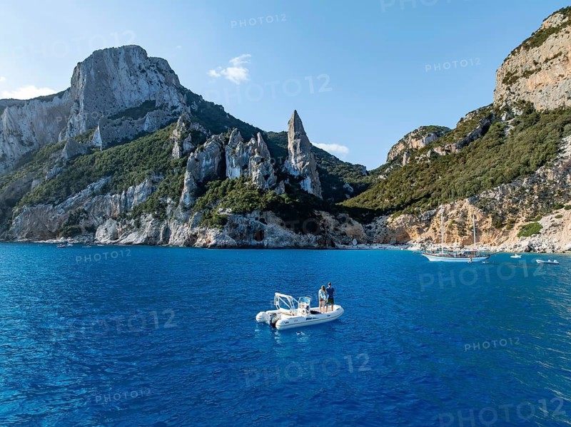 Couple on motorboat off picturesque rocky coast, cliffs with L'Aguglia pinnacle, blue sea and Cala Goloritzé beach, aerial view, Golfo di Orosei, Baunei, Sardinia, Italy