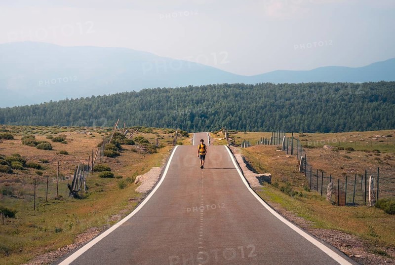A lone man wearing a backpack walks along a paved countryside road surrounded by lush greenery and distant mountains, symbolizing adventure and solitude in Guadarrama mountain range, Madrid