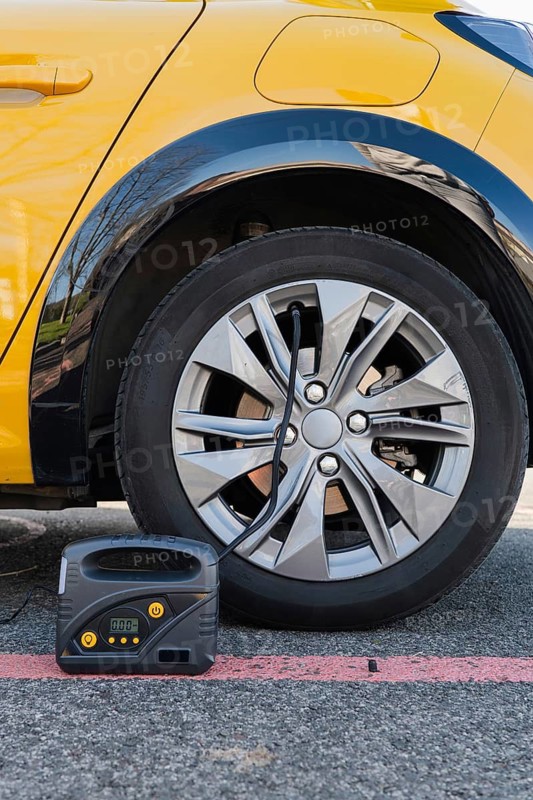 Close-up of an electric car's wheel with a portable tire inflator attached. The yellow vehicle is parked on an urban street, emphasizing modern automotive technology