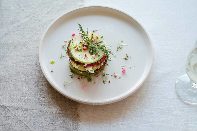 Top view of an artfully arranged gourmet salad featuring stacked slices of radish and cucumber, garnished with fresh dill and delicate flower petals, presented on a white plate with a glass of wine in the background