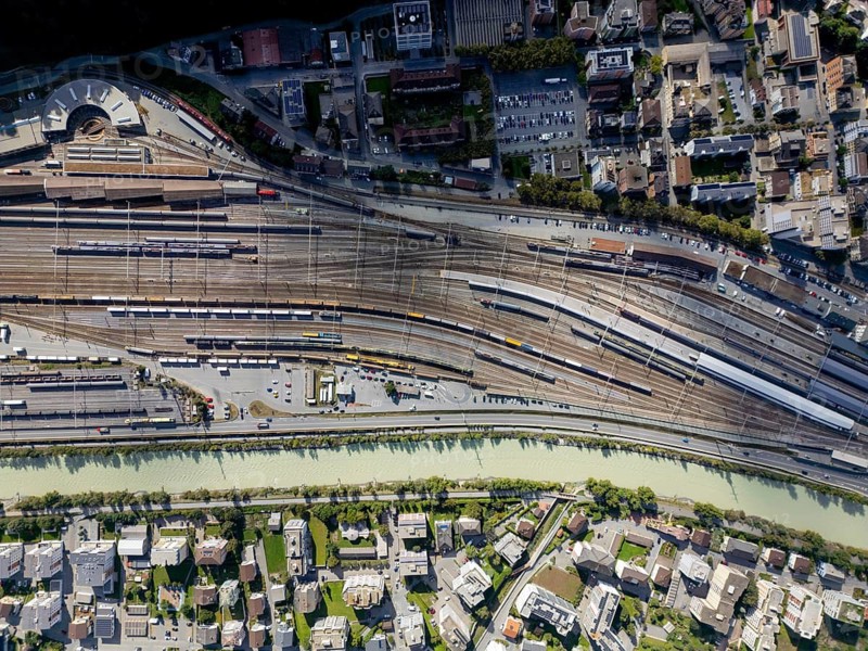 An aerial view of Brig railway station in Switzerland, showcasing a complex network of tracks and overhead wires, surrounded by urban buildings and lush greenery