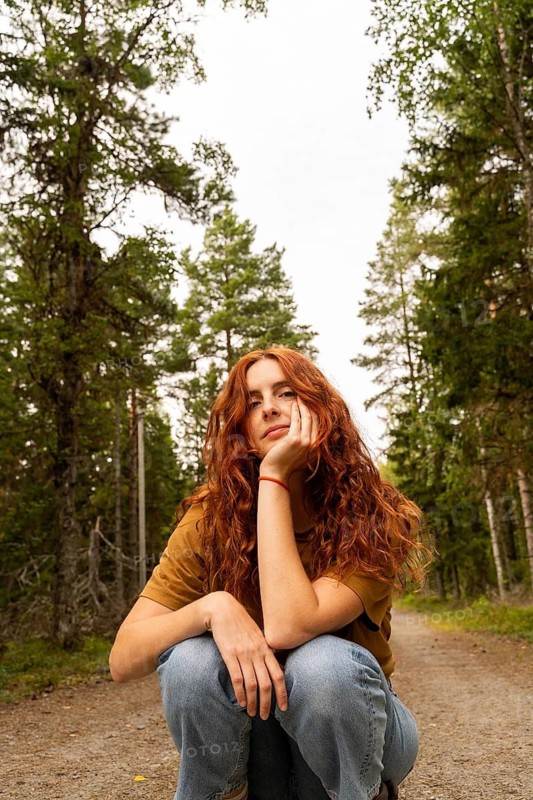 A young woman with red hair enjoys a peaceful moment in a Swedish forest, surrounded by tall trees. She is crouched on a dirt path, wearing a casual style