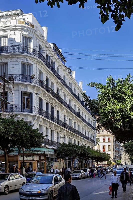 Lively street lined with white buildings, pedestrians and cars under a clear sky, The old town centre of Algiers in Algeria