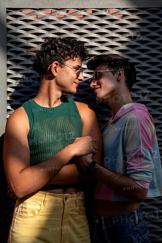 A joyful gay couple, wearing colorful outfits and sunglasses, shares a tender moment under the warm sunlight, leaning against a textured wall and embracing affectionately