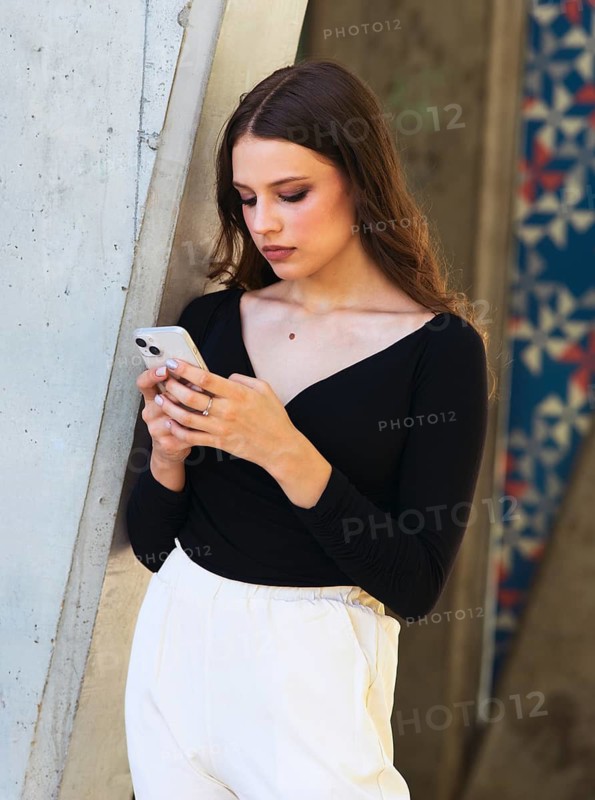 A woman in a black top and white pants leans against a concrete wall while using a smartphone. She appears focused and thoughtful, set against a colorful, urban background