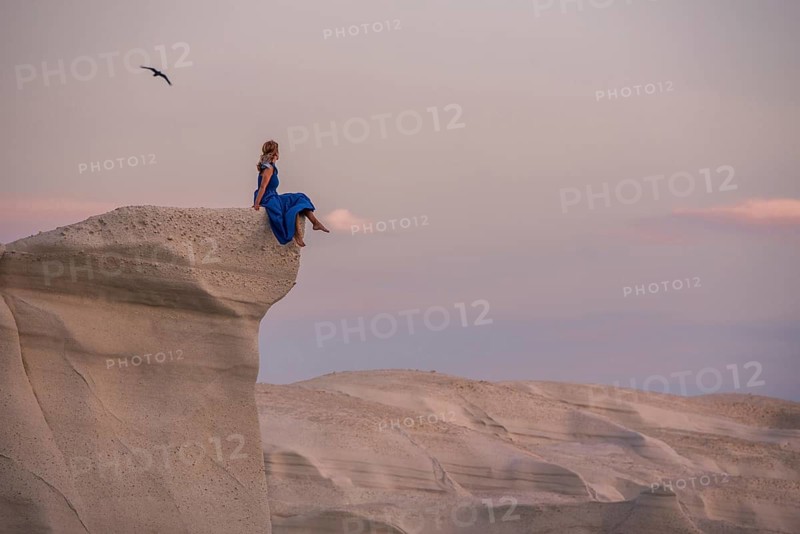 Unrecognizable woman wearing a blue dress perched on a high white rock formation at Sarakiniko on the island of Milos, Greece, during sunset