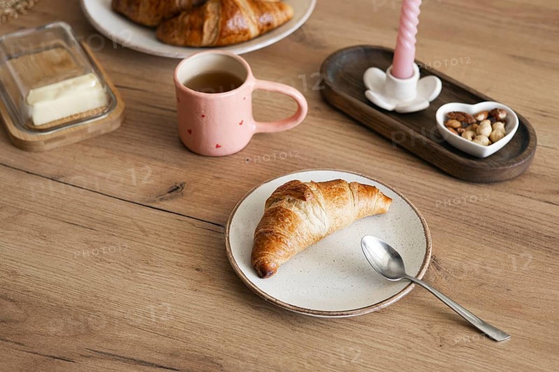 Delicious homemade croissants served on a rustic wooden table with a pink mug filled with coffee, and a dish of assorted nuts for a perfect breakfast setting
