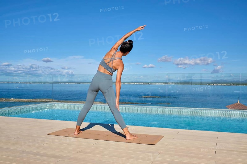 A woman practices yoga on a rooftop with a stunning ocean view. Dressed in blue workout attire, they perform a side stretch on a mat, embodying tranquility and balance