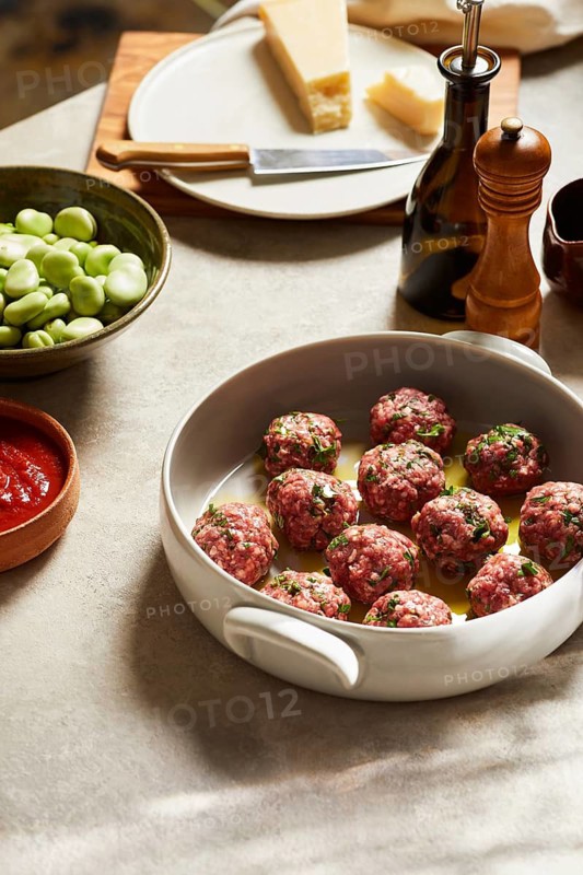 Raw meatballs seasoned with herbs in a white dish ready for cooking, surrounded by fresh ingredients like green beans, parmesan cheese, and tomato sauce on a kitchen counter