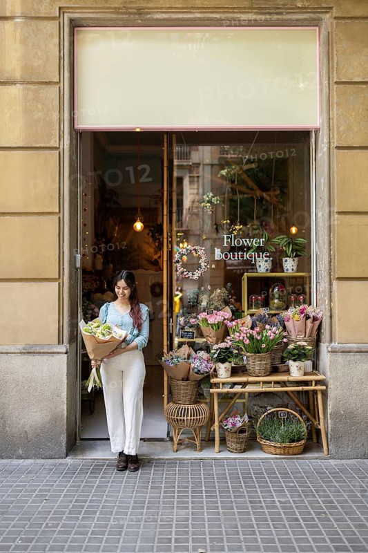 A woman stands at the entrance of a charming flower shop, holding a vibrant bouquet. Blooming flowers and lush plants surround the storefront, creating a welcoming and enchanting ambiance
