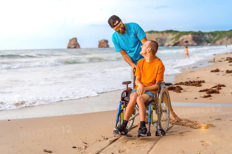 A disabled person in a wheelchair on the beach pushed by a friends by the sea, enjoying the summer