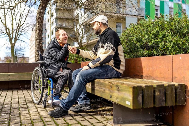 Two friends shaking hands while one sits in a wheelchair, both enjoying a sunny day together in the vibrant city park