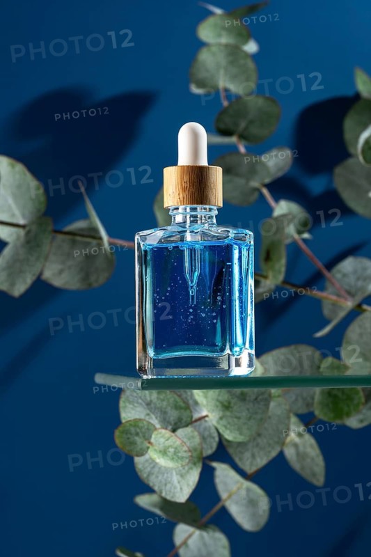 A blue cooling face serum for men, presented in a clear glass dropper bottle, elegantly surrounded by eucalyptus leaves against a deep blue background