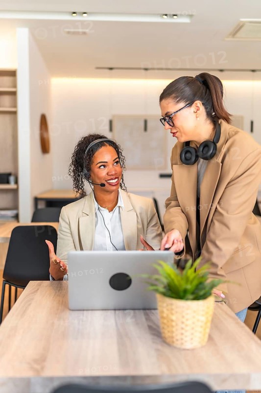 Two female customer service representatives are collaborating on a laptop, discussing work related matters in a modern office setting