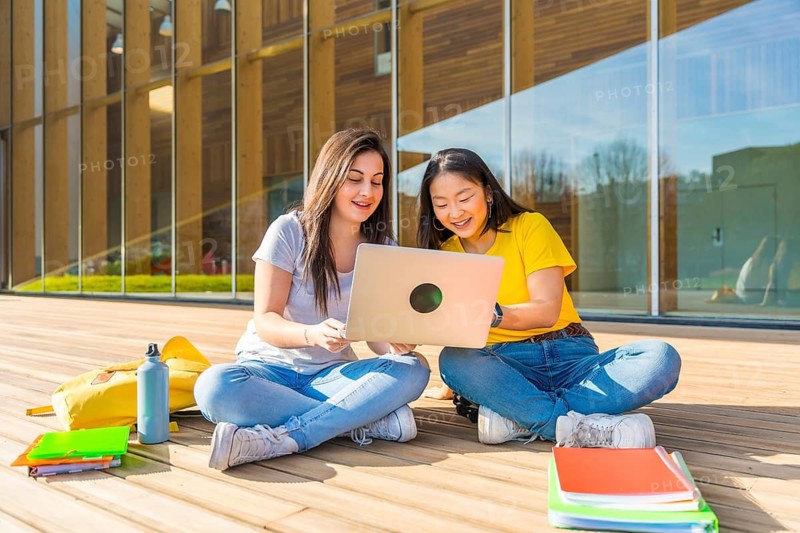Frontal photo of two multi-ethnic female friends using laptop sitting on the university campus