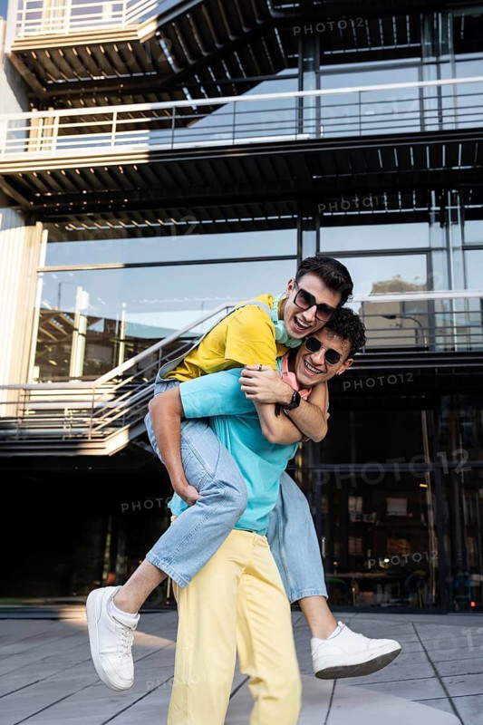 A happy gay couple shares a playful moment outdoors, with one partner giving the other a piggyback ride. They both wear colorful attire and sunglasses, set against a modern building