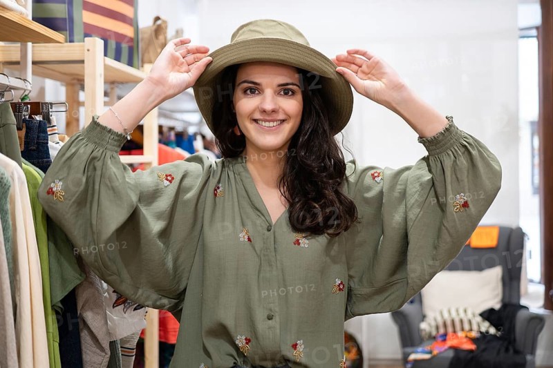 A woman smiles in a clothing store, wearing a green hat and embroidered dress. The setting is casual with vibrant clothes surrounding her