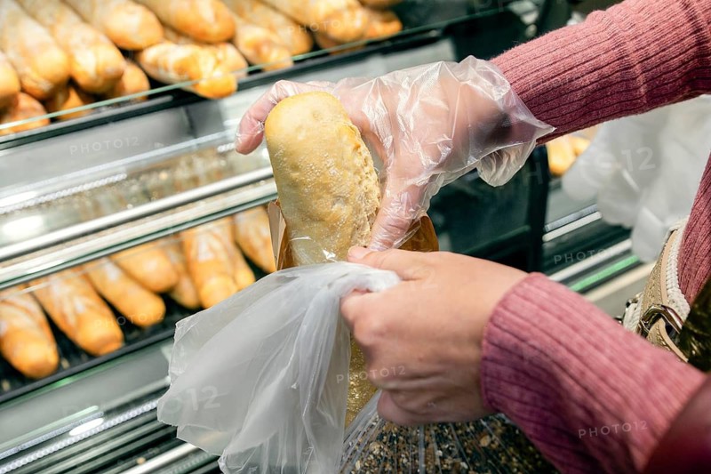 A woman wearing gloves selects a fresh loaf of bread from a bakery display at a supermarket. The scene captures everyday grocery shopping with hygiene precautions