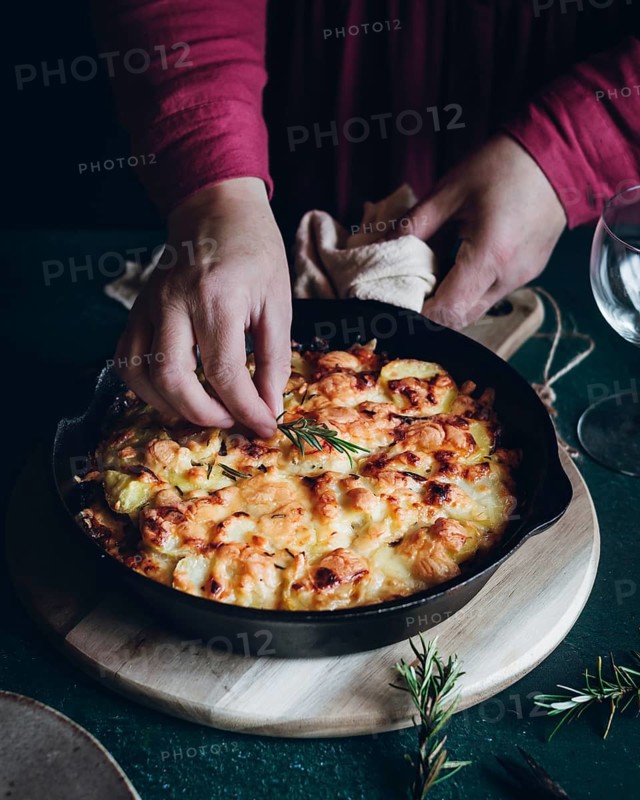 A close up of hands garnishing a cheesy potato gratin with fresh rosemary in a cast iron skillet. The scene evokes warmth and home cooked comfort