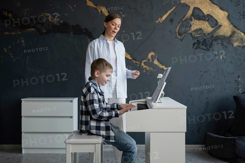 A young boy in a checkered shirt focuses on playing the piano in a modern room. His teacher, standing beside him, provides guidance and encouragement during the lesson