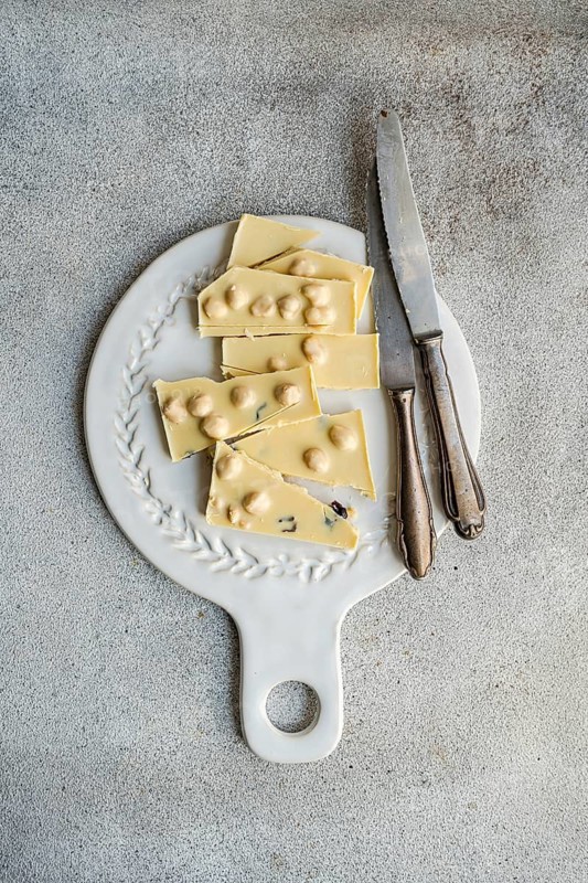 From above view of a white chocolate bar cut into pieces, placed on an ornate vintage cutting board with an old knife, showcasing a rustic, textured concrete background