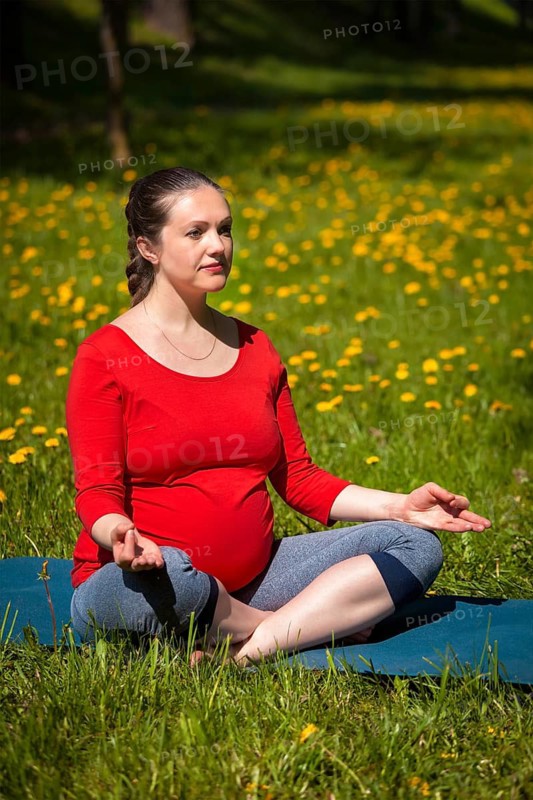 Pregnancy yoga exercise, pregnant woman doing asana Sukhasana easy yoga pose with chin mudra outdoors on grass lawn with dandelions in summer