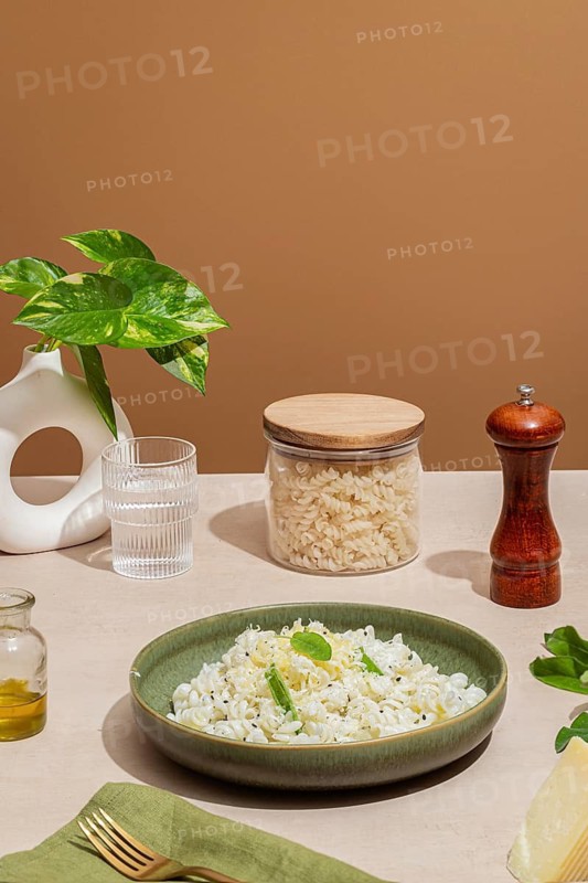 A beautifully styled table featuring a bowl of pasta garnished with herbs, surrounded by a jar of pasta, pepper mill, glass of water, olive oil, plant, and cutlery
