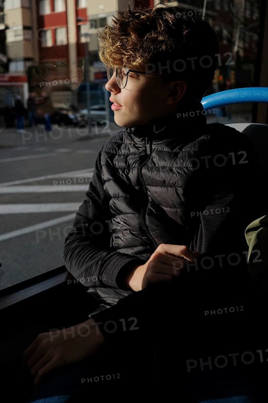 A young man with glasses sits on a bus, looking thoughtfully out the window. Sunlight highlights their face and reflection, creating a serene moment of contemplation