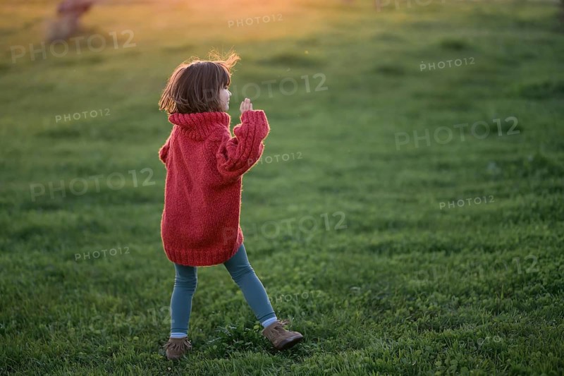 A young girl, wearing a cozy red sweater and blue pants, wanders through a lush green field during a golden autumn sunset, evoking a sense of warmth and curiosity