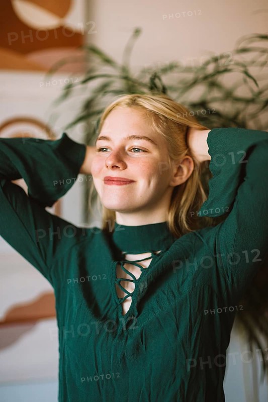 A teenager with a bright smile poses indoors, arms raised with confidence. The warm ambiance and greenery in the background create a relaxed atmosphere